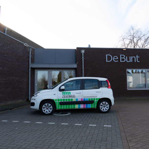 Voorzijde gebouw bloedafnamelocatie Gemeenschapshuis De Bunt in Someren. Met geparkeerde auto met logo Anna Ziekenhuis. Bij het Anna Ziekenhuis kun je op meerdere locaties in de regio bloed laten afnemen en lichaamsmateriaal zoals urine en ontlasting inleveren.