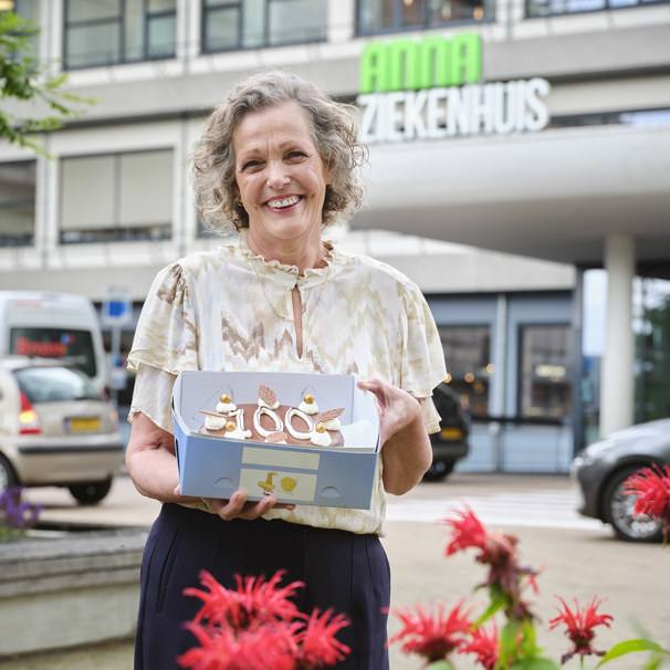 Marian staat voor de hoofdingang van het ziekenhuis en presenteert trots een taart met het cijfer 100 erop Marian nam voor haar 100e behandeling gebak mee voor de afdeling oncologie