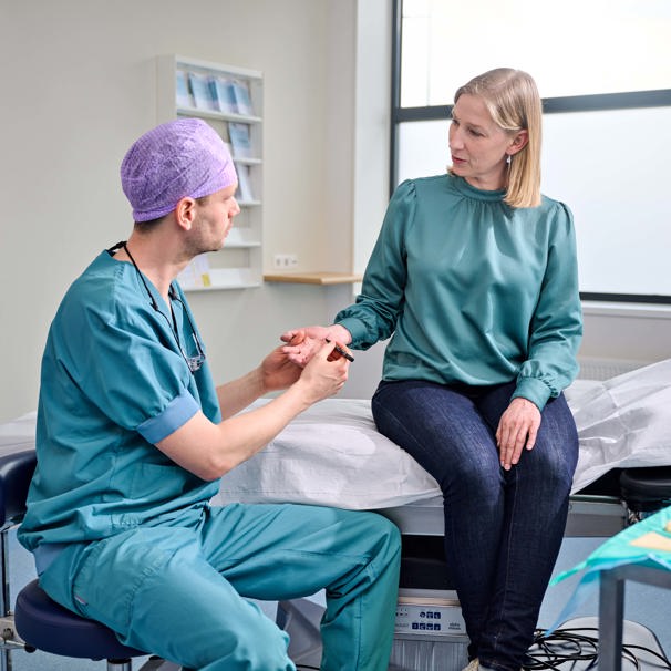 Dokter zit op stoel naast patiënt en voelt aan haar hand, terwijl patiënt naast hem op behandeltafel zit Plastische chirurgie Anna Ziekenhuis voor reconstructieve chirurgie, hand- en polschirurgie en cosmetische chirurgie