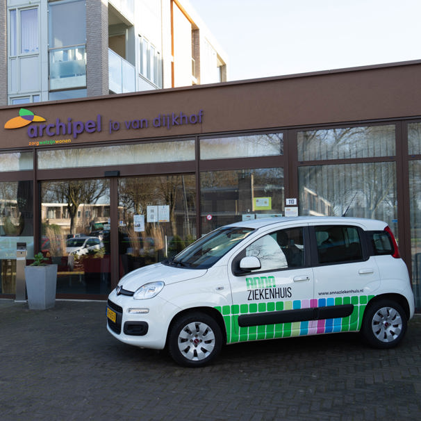 Voorzijde gebouw bloedafnamelocatie Gasterij Jo Van Dijkhof in Nuenen. Met geparkeerde auto met logo Anna Ziekenhuis. Bij het Anna Ziekenhuis kun je op meerdere locaties in de regio bloed laten afnemen en lichaamsmateriaal zoals urine en ontlasting inleveren.