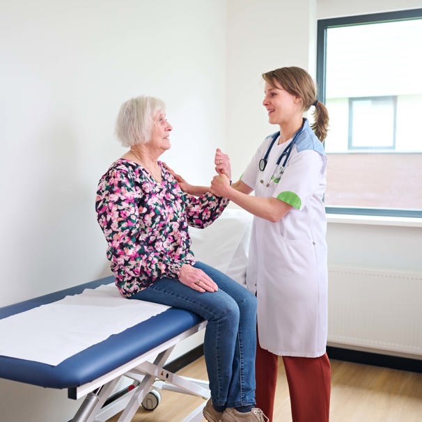 Patiënt zit op behandeltafel met haar arm omhoog, zorgverlener houdt de arm vast, terwijl ze met elkaar praten Bij geriatrie werken we aan het behoud en herstel van zelfstandigheid en kwaliteit van leven bij oudere mensen