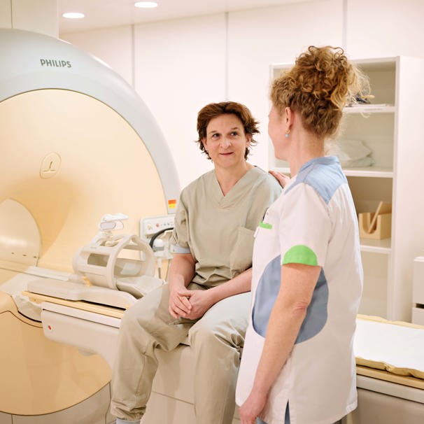 Patiënt zit bij MRI, verpleegkundige staat ernaast en ze zijn met elkaar in gesprek Radiologie voor onderzoeken van botten, weefsels en organen.