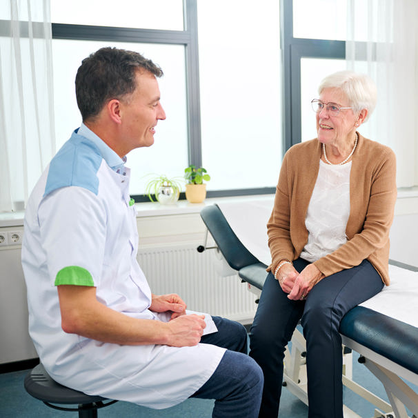 Dokter zit op stoel naast patiënt die op behandeltafel zit, ze kijken elkaar aan Orthopedie Anna Ziekenhuis kan helpen bij rugpijn, schouderklachten, heupklachten en kniepijn