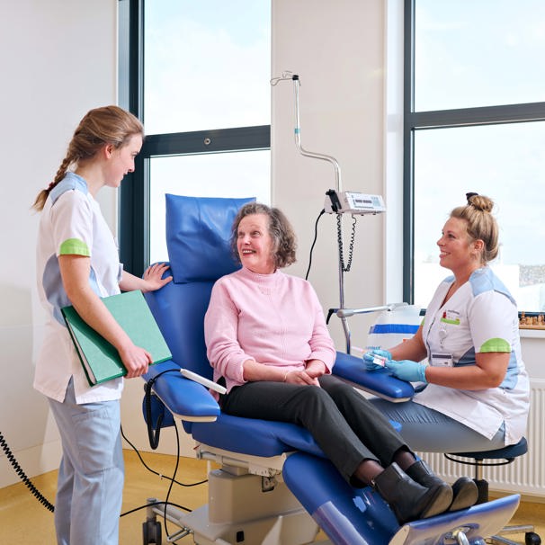 Patiënt zit op behandelstoel, aan haar linkerzijde zit een zorgverlener en aan haar rechterzijde staat een zorgverlener waarmee ze in gesprek is Oncologie voor onderzoek, behandeling en begeleiding bij alle vormen of voorstadia van kanker