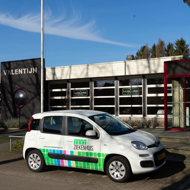 Voorzijde gebouw bloedafnamelocatie Dorpshuis Valentijn in Sterksel. Met geparkeerde auto met logo Anna Ziekenhuis. Bij het Anna Ziekenhuis kun je op meerdere locaties in de regio bloed laten afnemen en lichaamsmateriaal zoals urine en ontlasting inleveren.