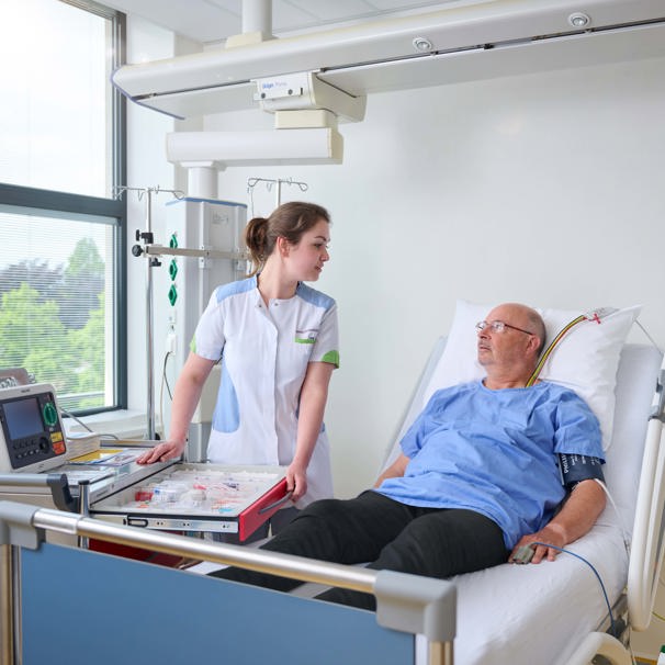 Patiënt ligt in ziekenhuisbed aan de hartbewaking, zorgverlener staat naast hem en ze kijken elkaar aan De IC-CCU-EHH biedt intensieve zorg en geavanceerde bewaking