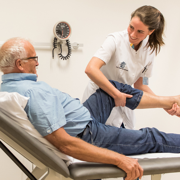 Patiënt ligt op behandeltafel, zorgverlener staat ernaast en houdt zijn been vast en omhoog, terwijl ze elkaar aankijken Reumatologie bij aandoeningen en klachten aan gewrichten, spieren en pezen die gepaard gaan met pijn en die je beperken in je bewegingen