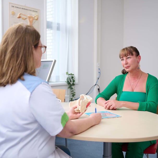 Dokter zit aan tafel met een medisch voorwerp in haar hand, patiënt zit aan andere kant van tafel en luistert Klachten bekkenbodem vrouwen