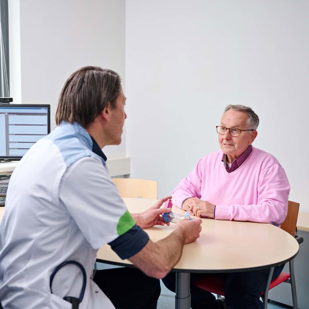 Patiënt zit op stoel aan tafel, aan de andere kant zit een dokter met een inhalator in zijn hand. Ze zijn in gesprek. Voor onderzoek en behandeling van aandoeningen aan de longen en luchtwegen